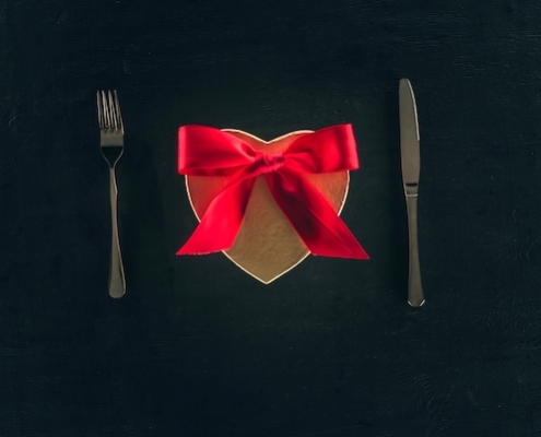The Best Edible Gifts to Stir Passion This Holiday Season 2 a place setting on a black table with a gold heart box tied with a red bow where the plate should be